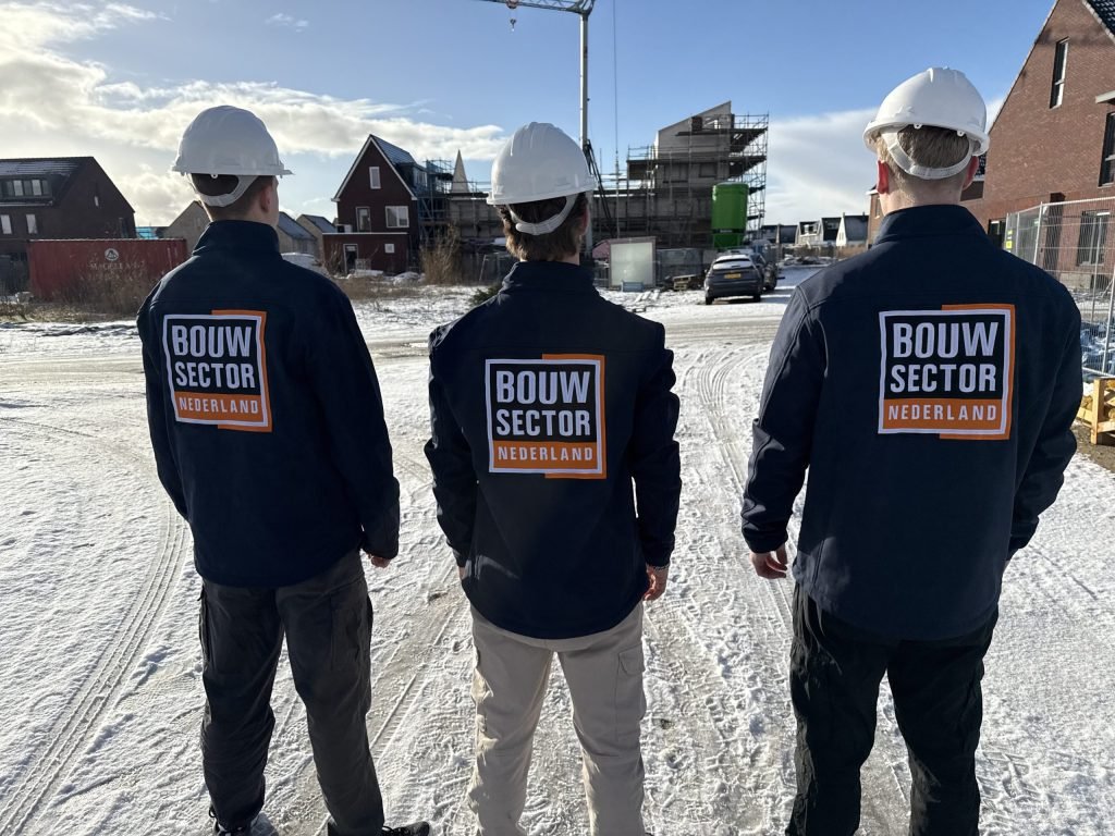 Drie vakmensen met witte helm en bedrijfsjas van Bouwsector Nederland op een besneeuwde nieuwbouwplaats